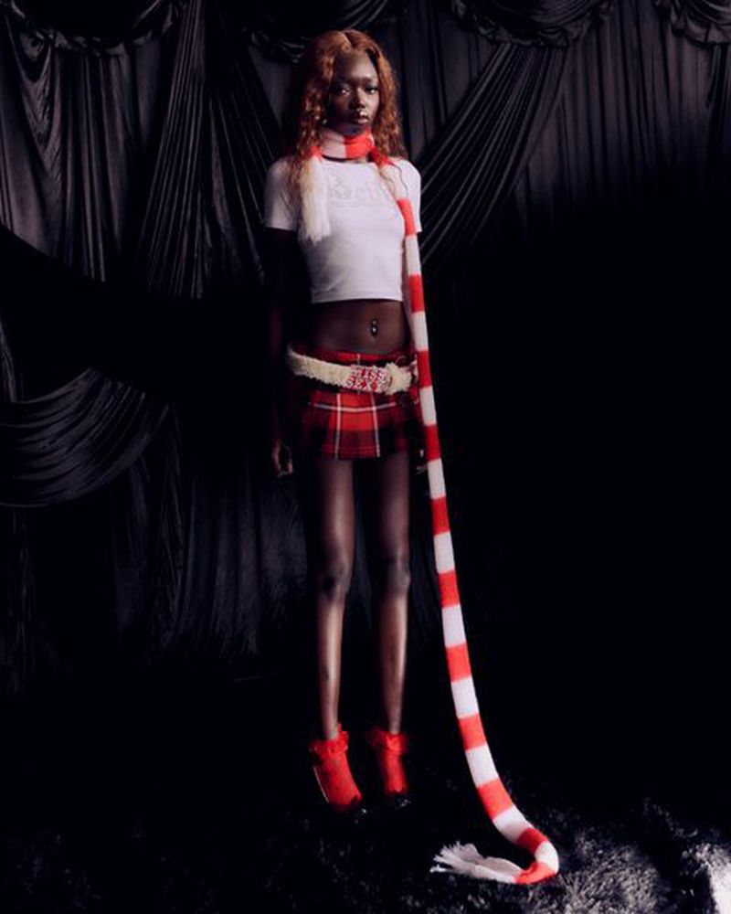 Young adult woman in a white crop top, red plaid skirt, red ankle boots, and a striped scarf, posing against a dark draped backdrop.