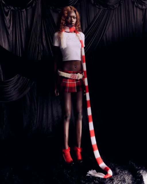 Young adult woman in a white crop top, red plaid skirt, red ankle boots, and a striped scarf, posing against a dark draped backdrop.