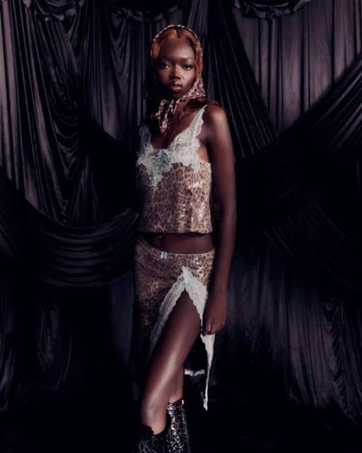 A woman with tan skin, adorned with a patterned scarf, poses in a matching sequined skirt and top against a dark, draped background.