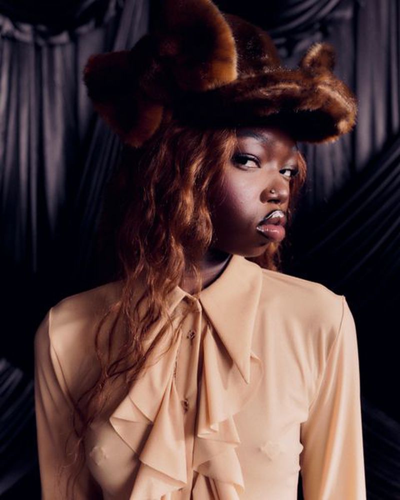 Portrait of a young Black woman wearing a fur hat and a beige blouse with ruffles, posing in a dimly lit indoor setting.