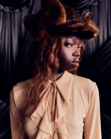 Portrait of a young Black woman wearing a fur hat and a beige blouse with ruffles, posing in a dimly lit indoor setting.