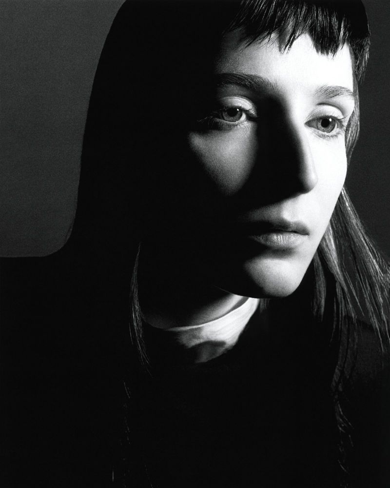 Black and white portrait of a young adult woman with dark hair, dramatic lighting, and a neutral facial expression in a photo studio.