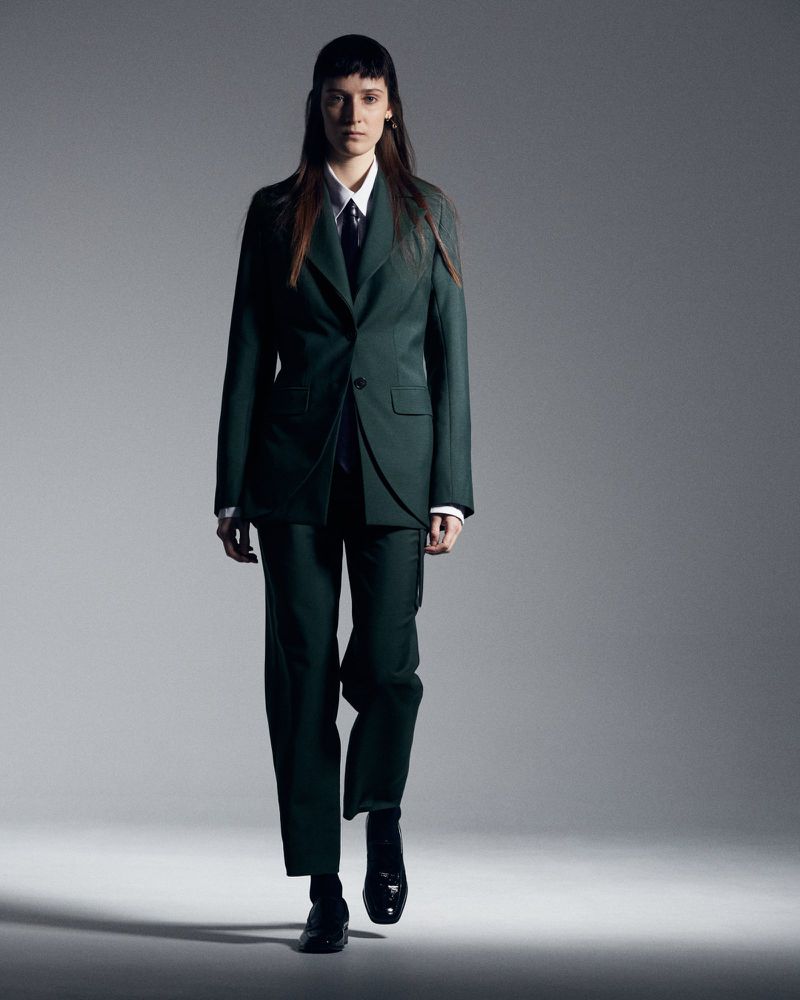 Fashionable adult female walks confidently in a stylish, dark-green pantsuit, white dress shirt, black tie, and black shoes against a gray backdrop.