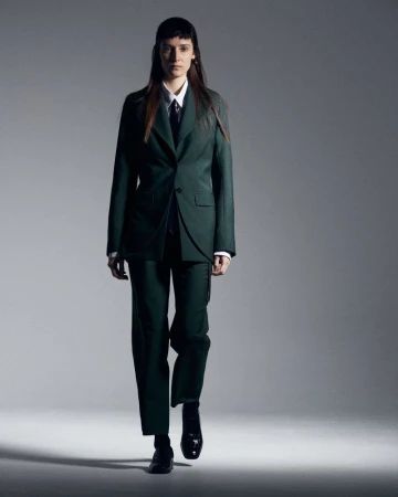 Fashionable adult female walks confidently in a stylish, dark-green pantsuit, white dress shirt, black tie, and black shoes against a gray backdrop.