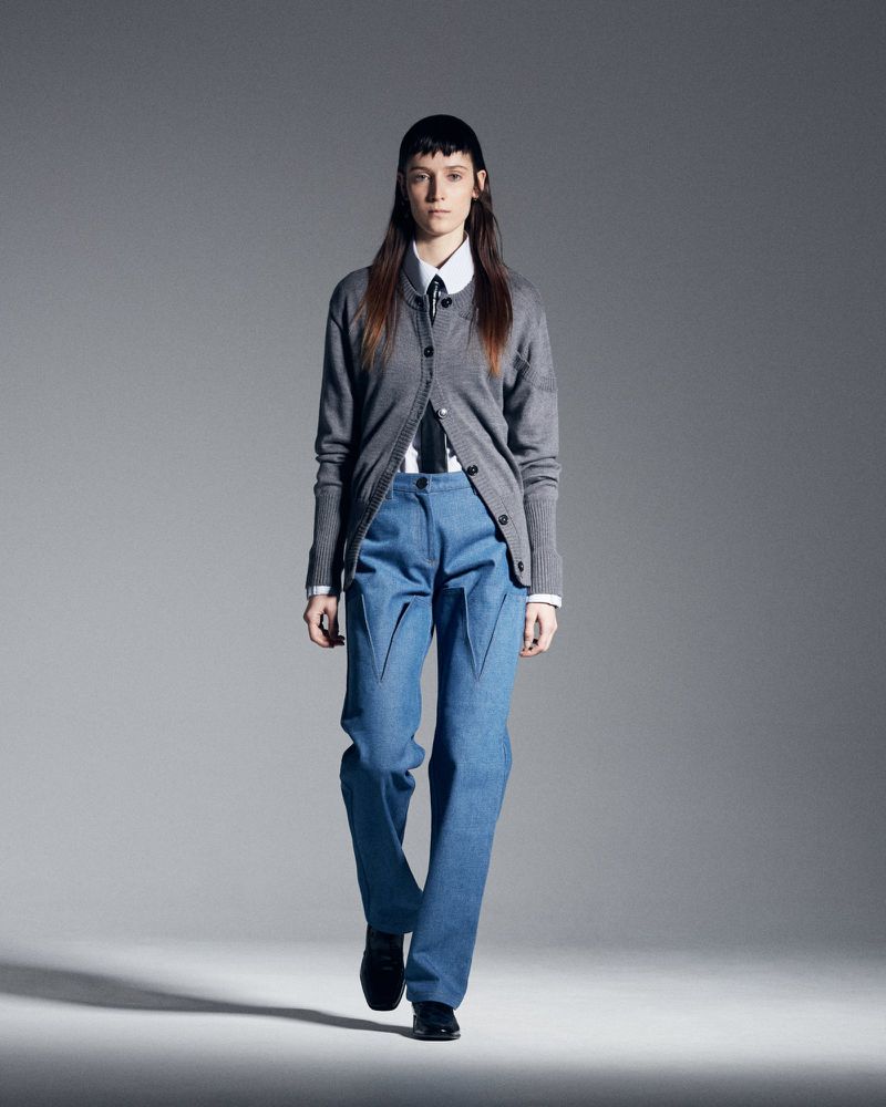 Runway fashion presentation shows a female adult model in a grey cardigan, white shirt, blue jeans, and black dress shoes. Studio lighting.