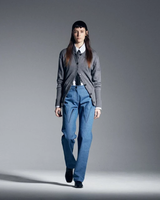 Runway fashion presentation shows a female adult model in a grey cardigan, white shirt, blue jeans, and black dress shoes. Studio lighting.