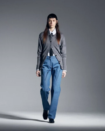 Runway fashion presentation shows a female adult model in a grey cardigan, white shirt, blue jeans, and black dress shoes. Studio lighting.
