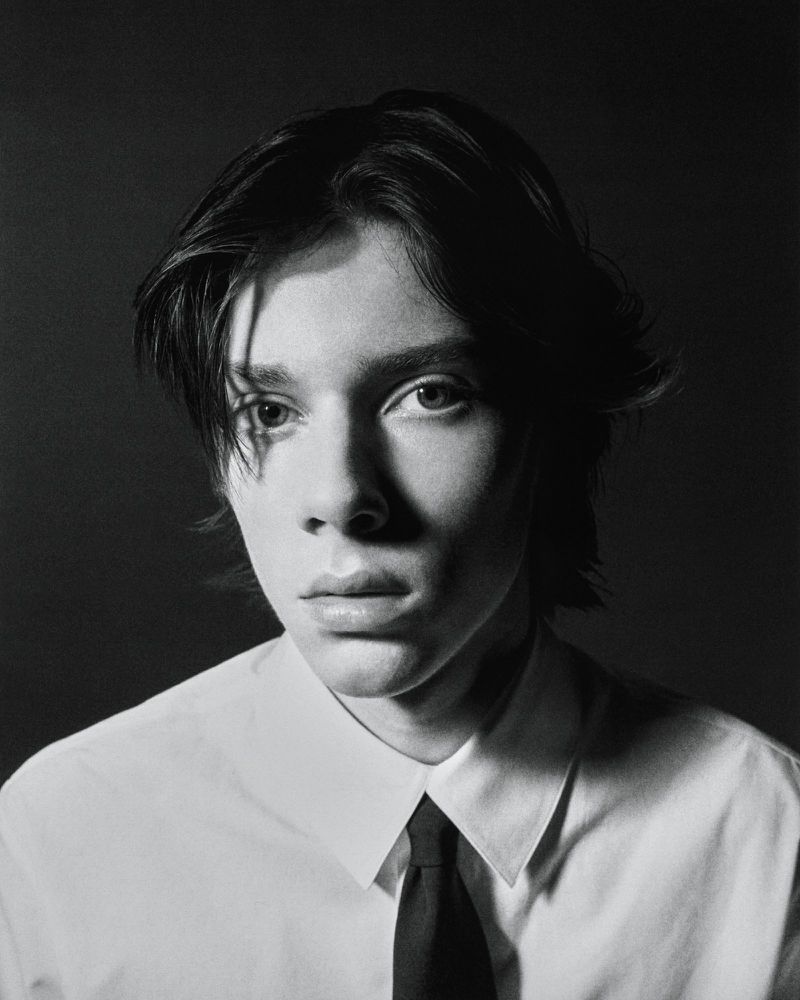 Monochrome portrait of a young adult with dark hair wearing a collared shirt and tie against a dark background.