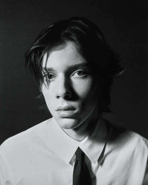 Monochrome portrait of a young adult with dark hair wearing a collared shirt and tie against a dark background.