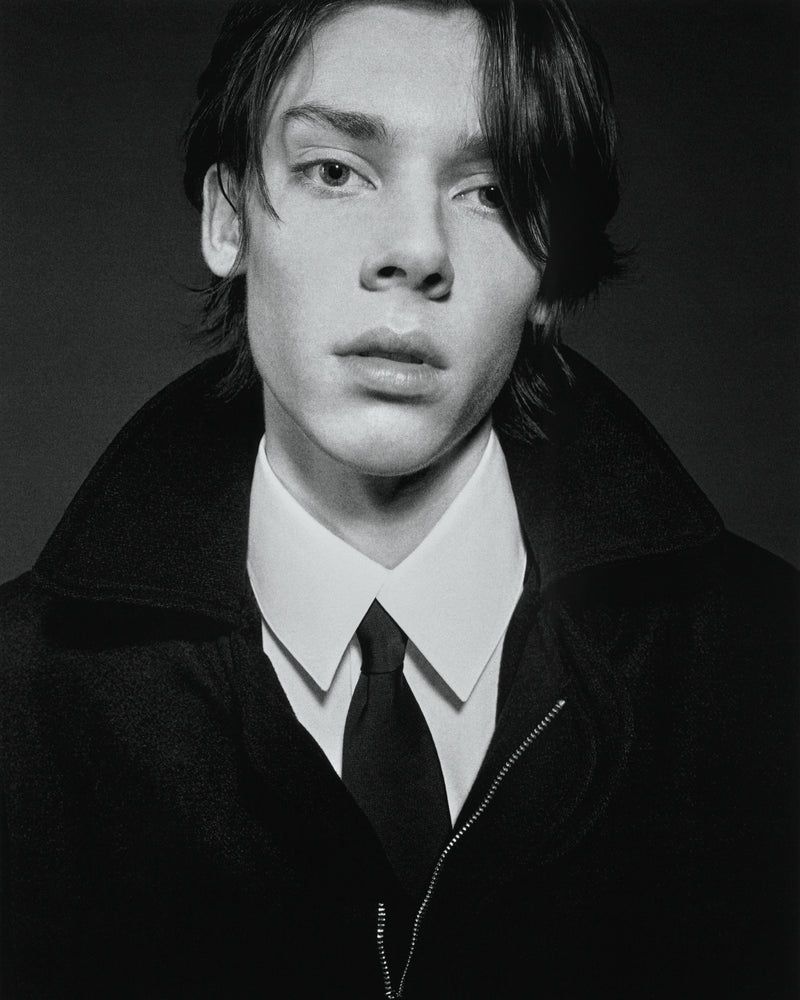 Portrait of a young adult male wearing a dark overcoat, white collared shirt, and a dark tie in a high-contrast studio shot.