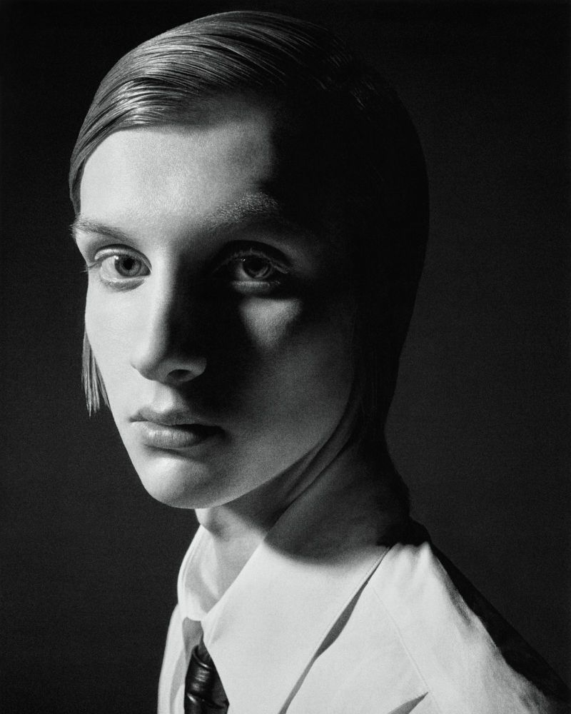 Black and white portrait of an adult woman with slicked-back short hair, wearing a white collared shirt and black tie, against a dark background.