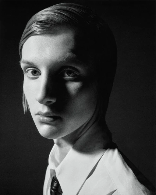 Black and white portrait of an adult woman with slicked-back short hair, wearing a white collared shirt and black tie, against a dark background.
