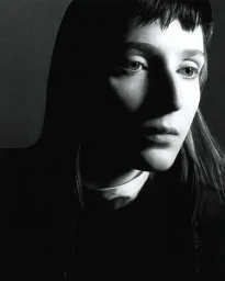 Black and white close-up portrait of a woman with short micro bangs illuminated by dramatic high-contrast shadow and light.