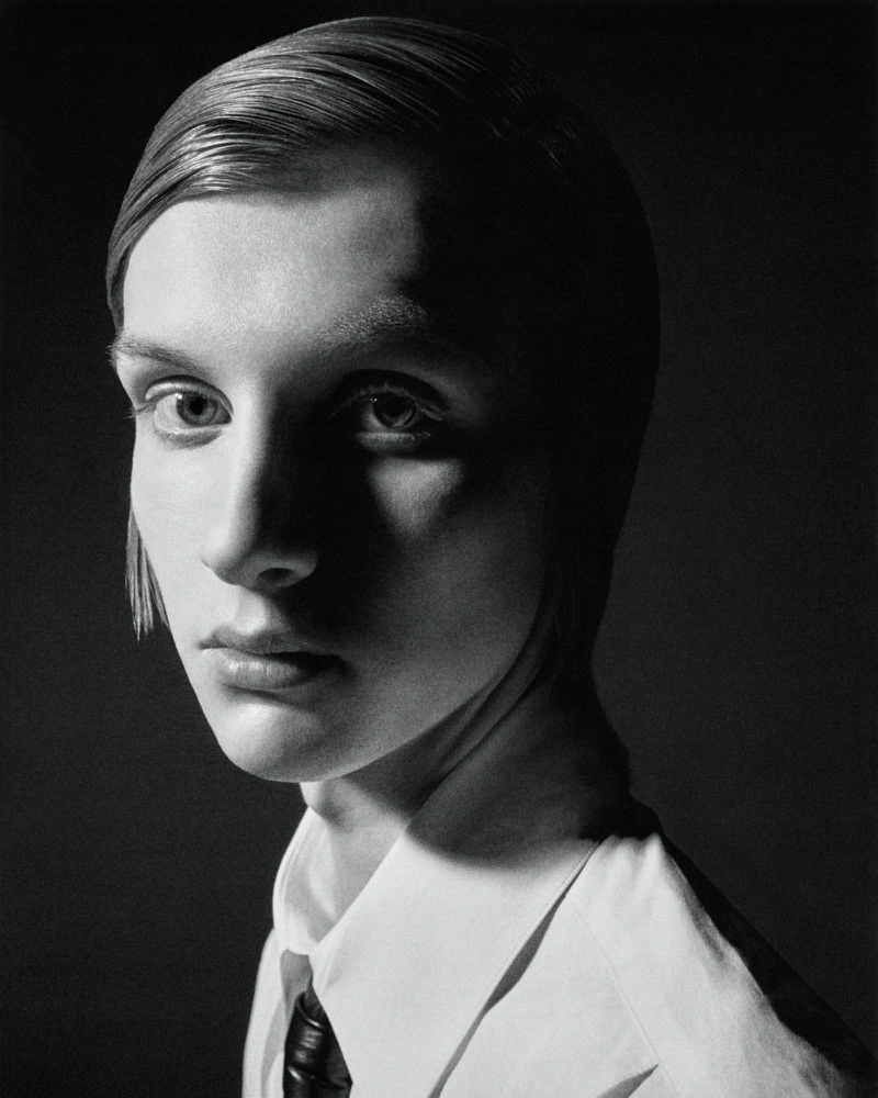 Black and white portrait of a model with slicked hair wearing a white collared shirt and dark tie against a black studio background.