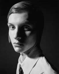Black and white portrait of a model with slicked hair wearing a white collared shirt and dark tie against a black studio background.