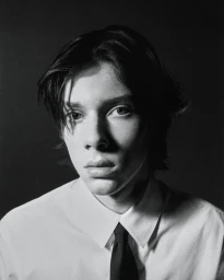 Black and white portrait of a young man wearing a white collared shirt and black tie against a dark background.