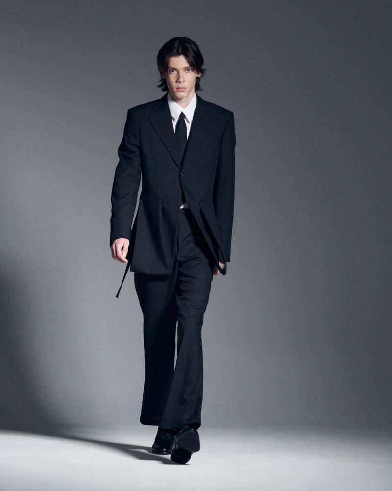 Model in black suit and white shirt walking against a grey seamless studio background.