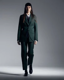 Model wearing a dark green tailored suit with a white shirt and dark tie against a grey seamless studio background.