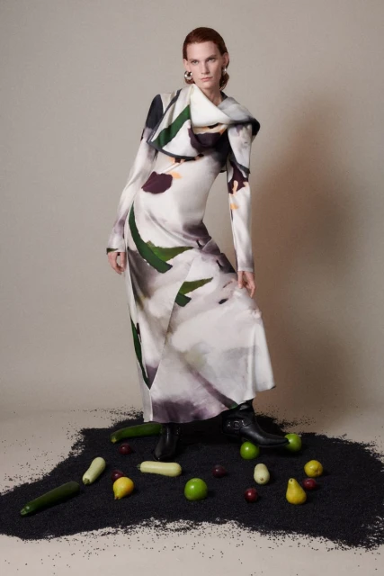 Fashion model in a long dress with a print, standing on a bed of dark material, surrounded by fresh fruit and vegetables in a photo studio.