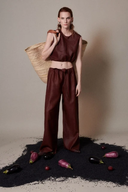 A slender woman wearing a red-brown two-piece outfit stands on a bed of black granules with eggplants, in a studio setting.