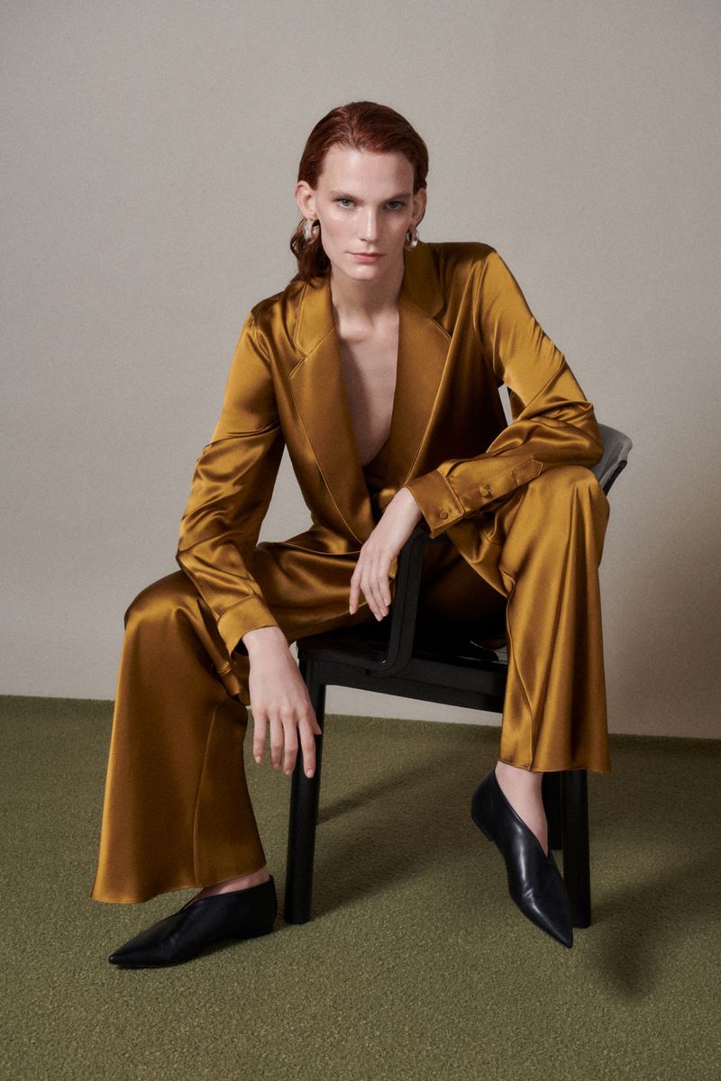 Adult female with red hair poses in a gold satin pantsuit on a black chair, with a neutral studio background and olive green carpet.