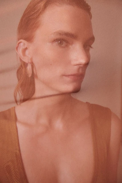Portrait of a fair-skinned adult with short, wavy hair, wearing a sleeveless top and earrings, and posing behind a net filter.