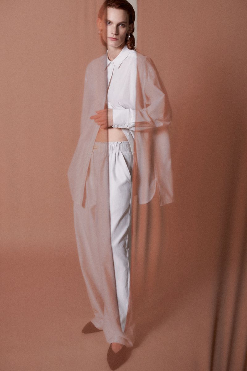 A blurry and layered fashion portrait features a tall person in a white shirt and pants, against a peach-toned backdrop in a studio setting.