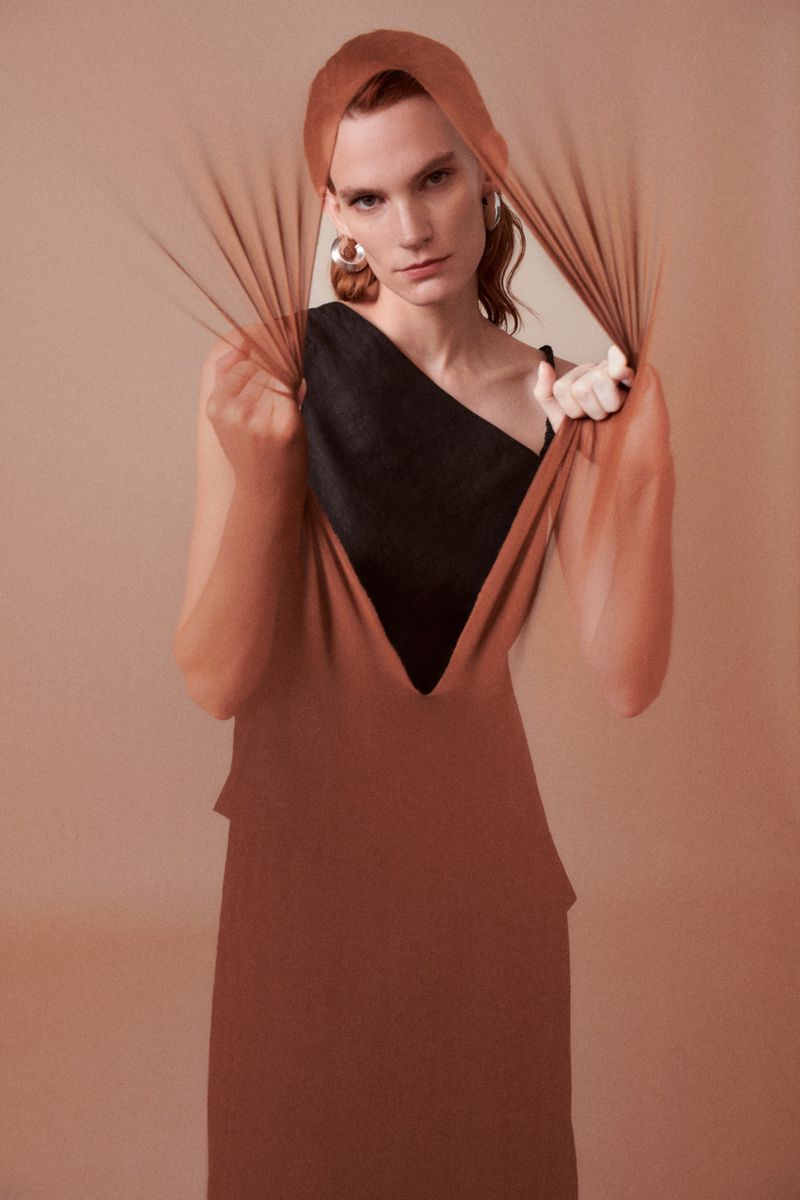 A model wearing a copper-colored dress and head covering, is seen indoors, framed by sheer fabric against a diffused peach background.
