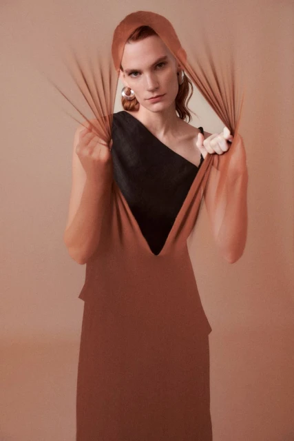 A model wearing a copper-colored dress and head covering, is seen indoors, framed by sheer fabric against a diffused peach background.