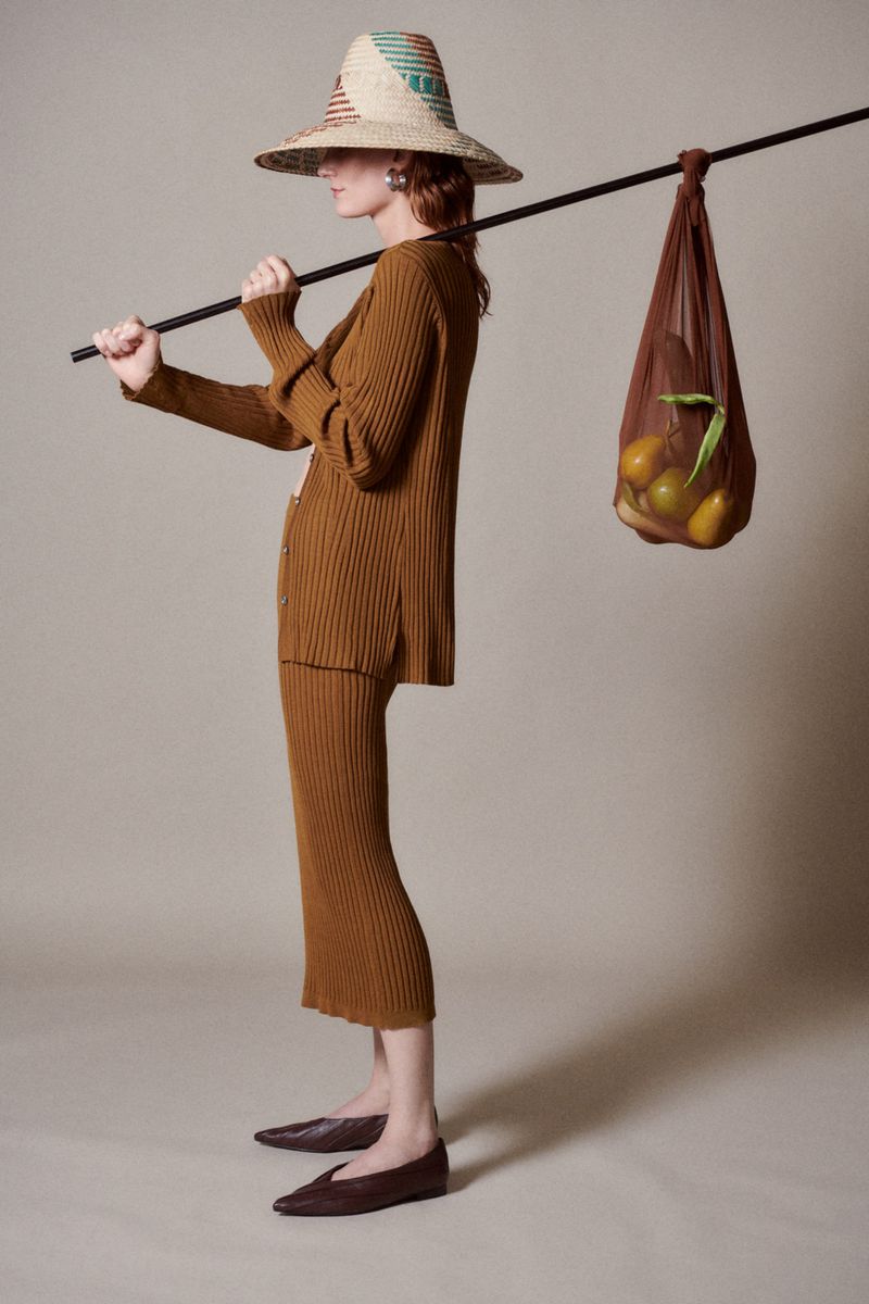 A young adult female in a brown knit skirt and cardigan carries a straw hat and a net bag full of oranges, studio lighting.