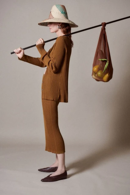 A young adult female in a brown knit skirt and cardigan carries a straw hat and a net bag full of oranges, studio lighting.