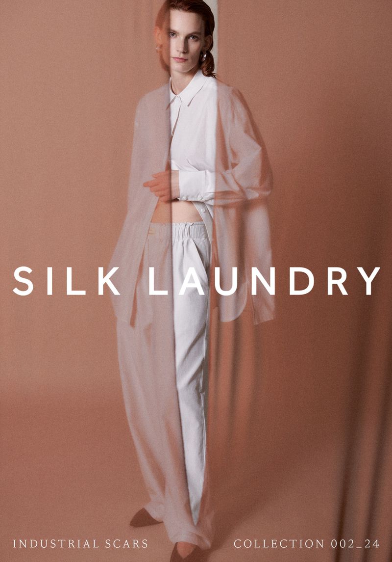 Fashion editorial shot of a fair-skinned woman in white pants and white top with a sheer beige outer layer against a beige backdrop.
