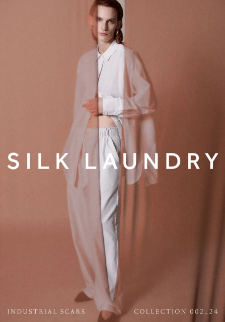 Fashion editorial shot of a fair-skinned woman in white pants and white top with a sheer beige outer layer against a beige backdrop.