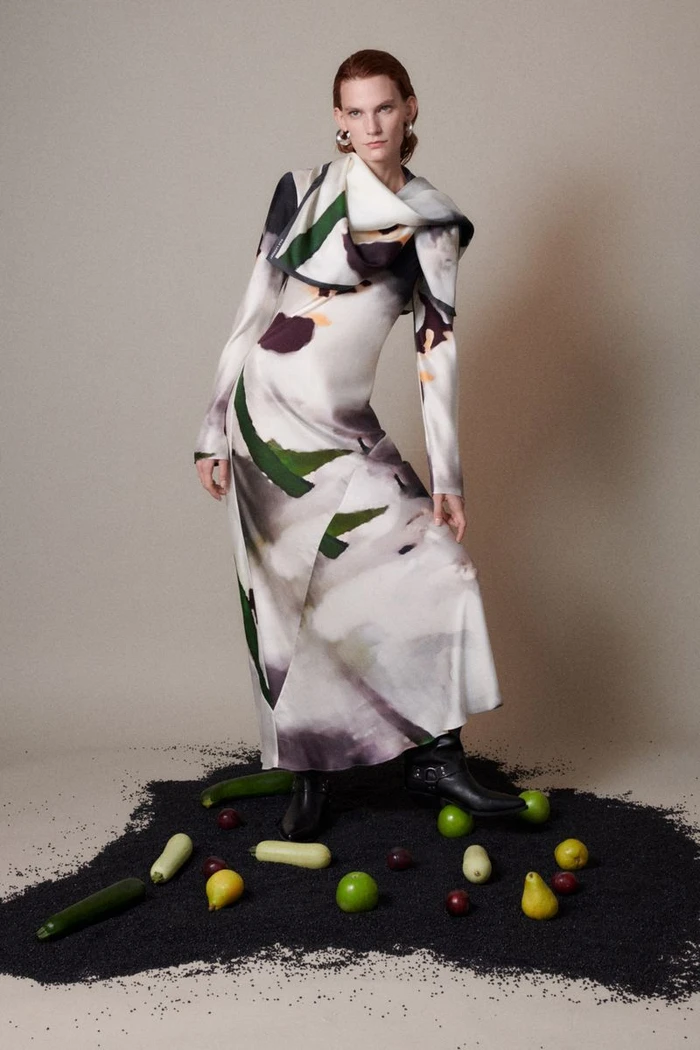 Model Lena Hardt in a printed maxi dress standing on black grain among scattered fruit against a grey studio backdrop.