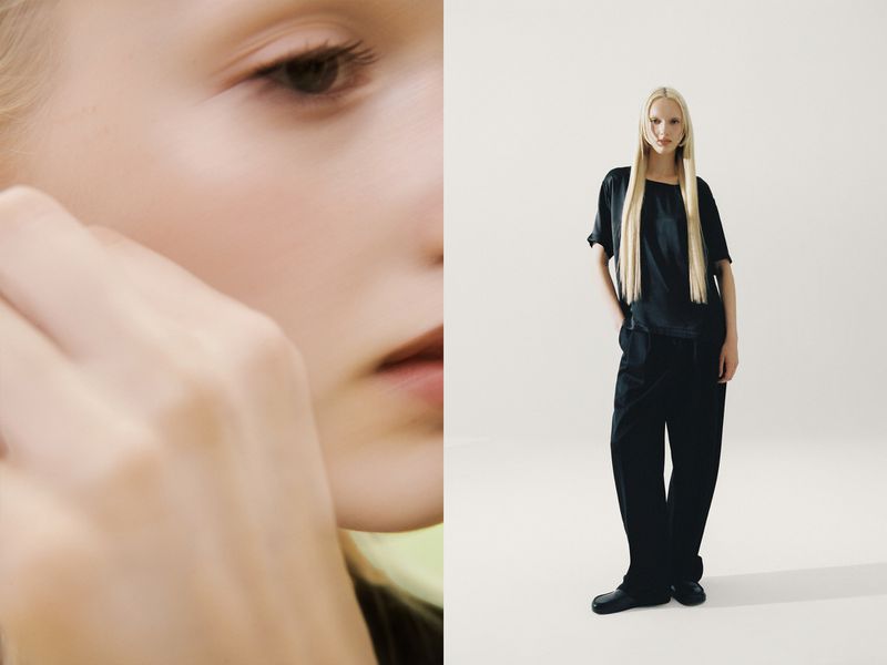 Fashion shot: a fair-skinned, light-haired young adult woman wears a black top and pants against a minimalist white backdrop in a studio.