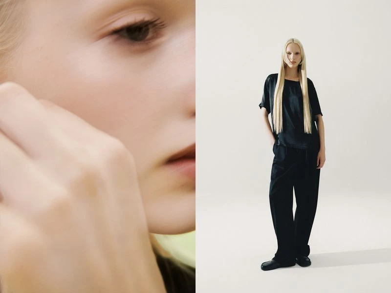 Fashion shot: a fair-skinned, light-haired young adult woman wears a black top and pants against a minimalist white backdrop in a studio.