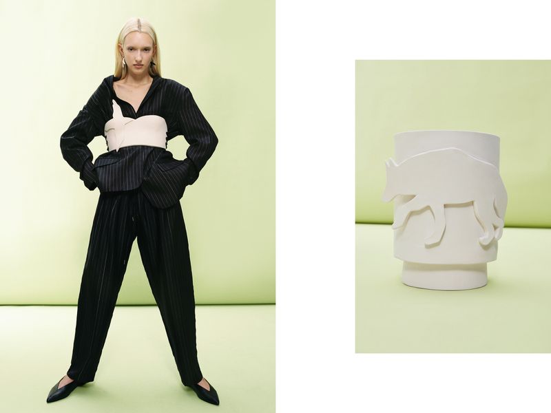Fashion shot: Young, fair-skinned woman in pinstripe suit and cream top stands in front of pale green background, vase with animal design.