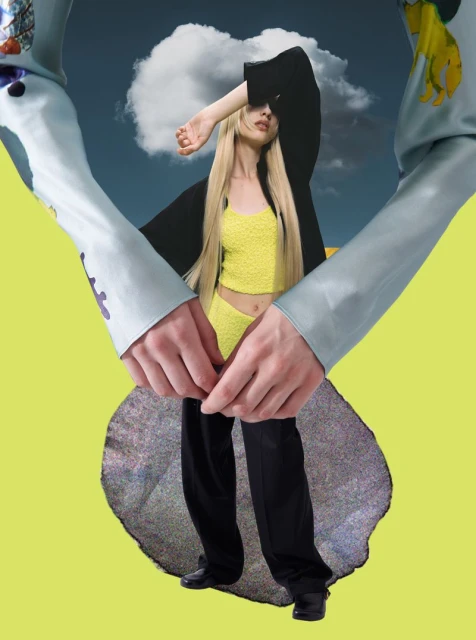 Woman in yellow crop top and black pants poses with hands and arms creating a frame against a blue sky with clouds and yellow backdrop.