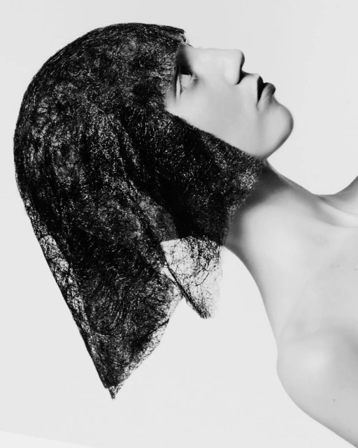 Monochrome portrait of an adult woman with bare shoulders, looking up and wearing a lace head covering against a light grey background.