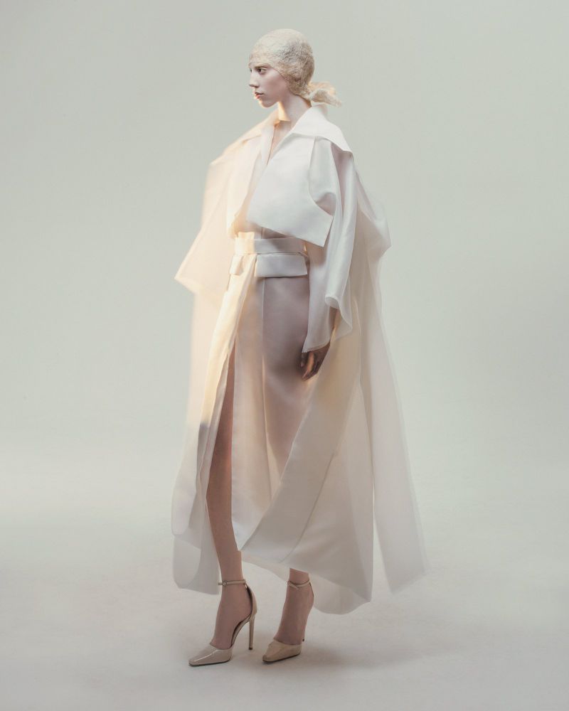 Model posing in a sculptural draped white layered coat and high-slit wrap skirt with stiletto heels against a neutral studio backdrop.