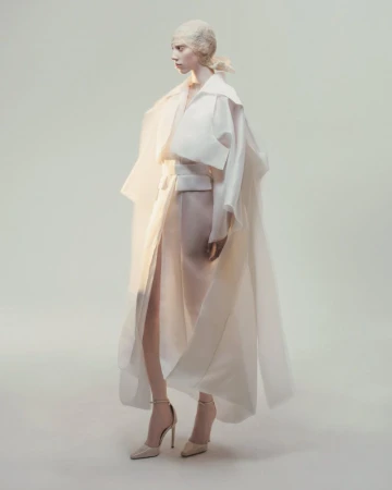 Model posing in a sculptural draped white layered coat and high-slit wrap skirt with stiletto heels against a neutral studio backdrop.