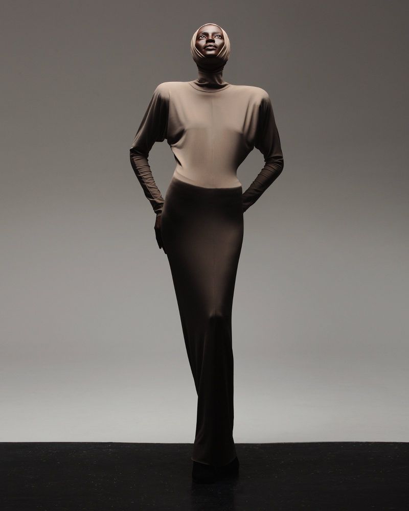 A slender adult woman in a long tan dress with a head wrap poses gracefully against a graduated grey background in a studio setting.
