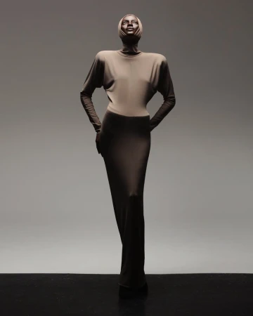 A slender adult woman in a long tan dress with a head wrap poses gracefully against a graduated grey background in a studio setting.