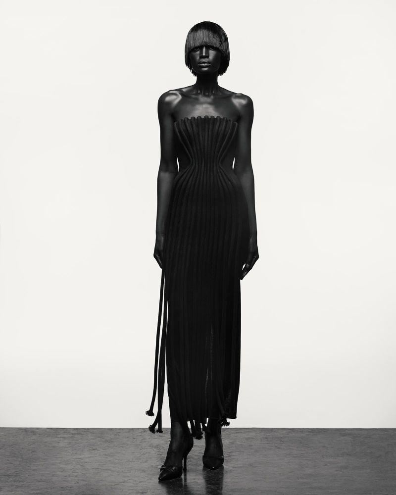 A black-clad fashion model presents a strapless, pleated long dress with matching high heels in a full shot against a white backdrop.