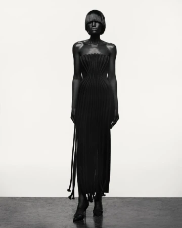 A black-clad fashion model presents a strapless, pleated long dress with matching high heels in a full shot against a white backdrop.