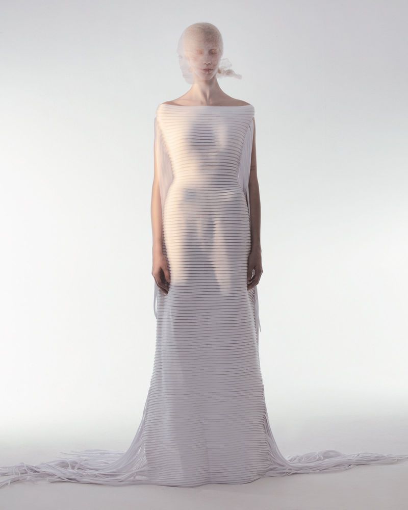 Woman in a pleated gown with a fabric veil, standing in a light studio setting, showcasing fashion and artistic photography.