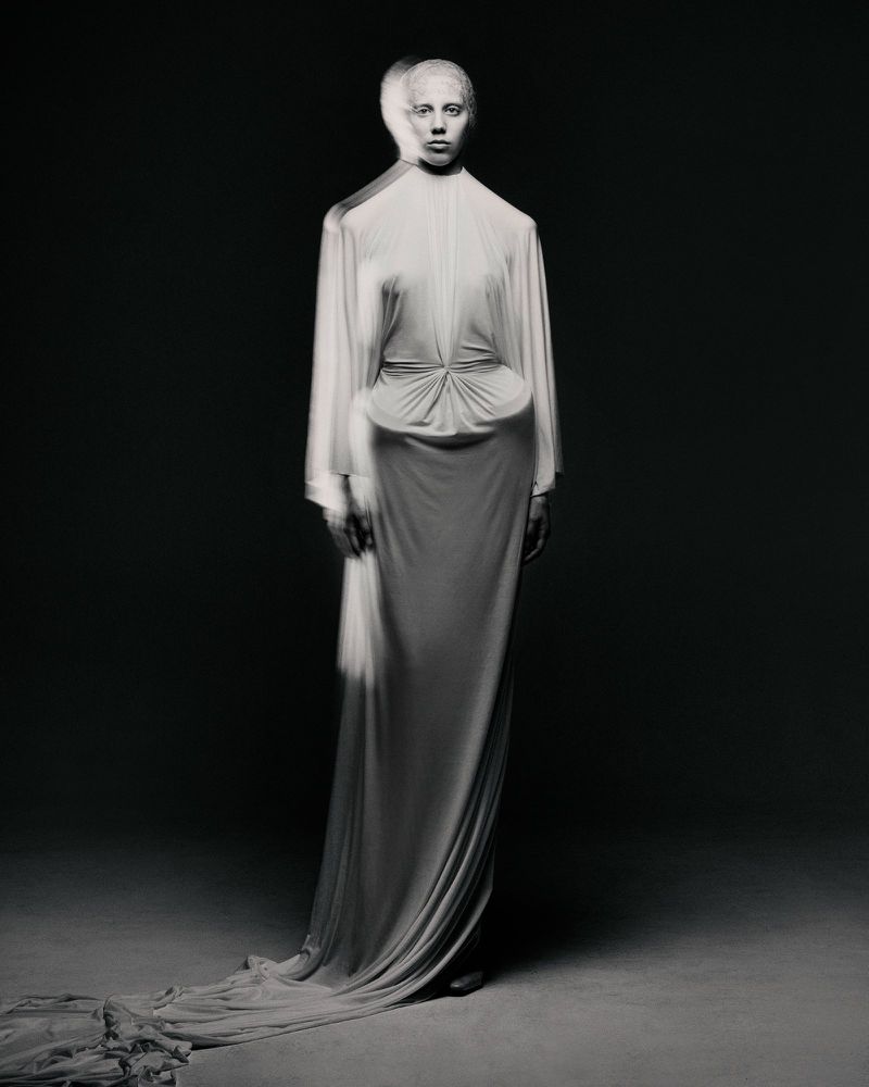 A tall slender model in a long flowing white dress stands in a photo studio against a gradient grey background with soft directional lighting.