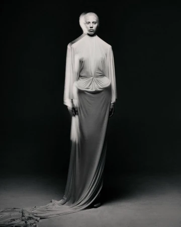 A tall slender model in a long flowing white dress stands in a photo studio against a gradient grey background with soft directional lighting.
