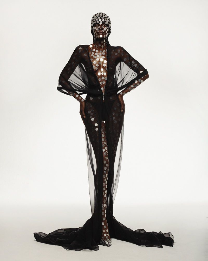 Model wearing a sheer black floor-length gown adorned with metallic circular discs against a white studio background.