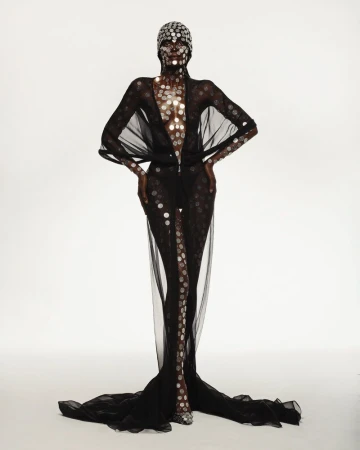 Model wearing a sheer black floor-length gown adorned with metallic circular discs against a white studio background.